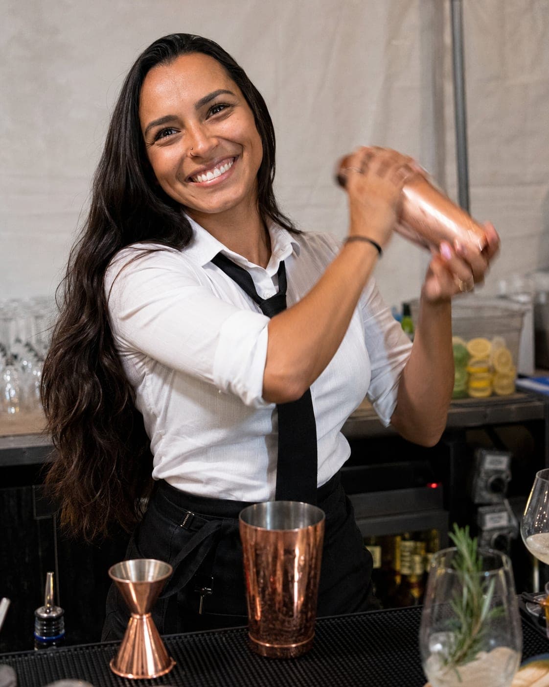 A professional bartender in a white shirt and black tie serving drinks behind a bar at a tented outdoor event